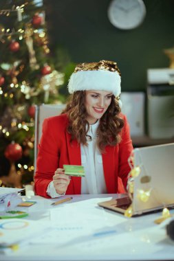 Noel zamanı. Gülümseyen, Noel Baba şapkalı ve kırmızı ceketli modern iş kadını. Laptop ve kredi kartıyla modern yeşil ofis ve Noel ağacı..