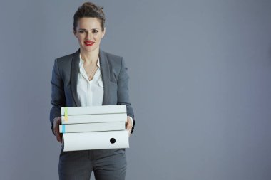 Gri arkaplanda dosyalar bulunan gri takım elbiseli, güler yüzlü orta yaşlı kadın çalışan..