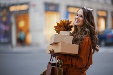 Merhaba Ekim. Turuncu trençkotlu, paketli, alışveriş torbalı ve sonbahar sarısı yapraklı gülümseyen genç kadın..