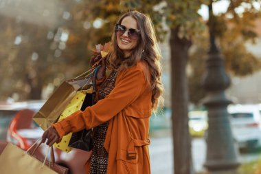 Merhaba Eylül. Şehirde alışveriş torbaları ve sonbahar sarısı yaprakları olan kahverengi trençkotlu mutlu şık bir kadın..