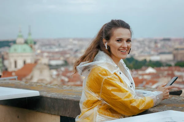 happy stylish traveller woman in yellow blouse and raincoat in Prague Czech Republic with headphones using smartphone against city panorama.