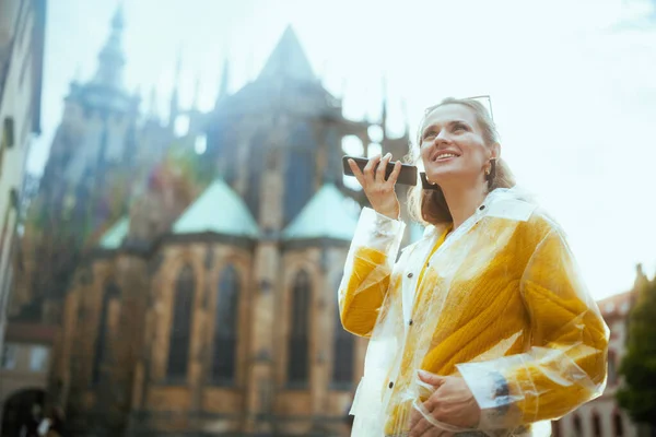 happy trendy solo tourist woman in yellow blouse and raincoat in Prague Czech Republic exploring attractions, listening to an audio guide on smartphone app and using a smartphone.