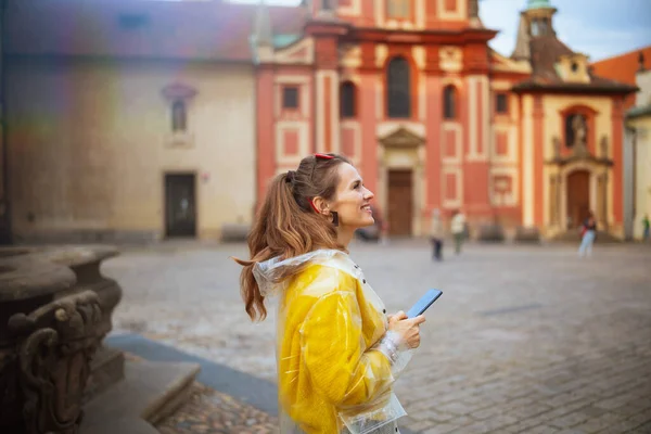 smiling modern woman in yellow blouse and raincoat in Prague Czech Republic with smartphone walking.