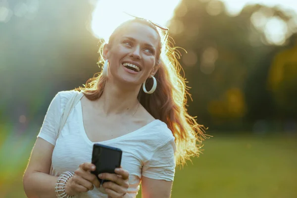 夏天的时候城市公园的草地上 穿着白衬衫的快乐中年妇女正在使用智能手机应用程序 — 图库照片