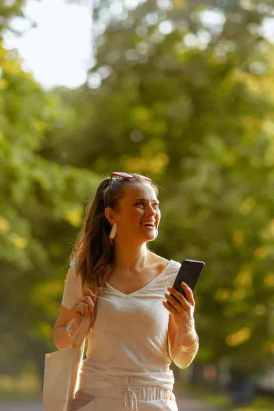 夏天的时候穿着白衬衫 头戴手提包 手持智能手机 走在城市公园里的时髦女人 — 图库照片