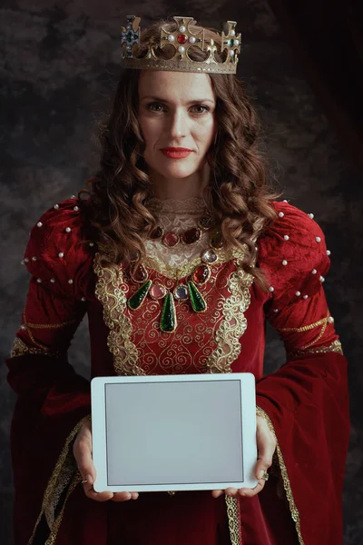 stock image medieval queen in red dress with crown showing tablet PC blank screen on dark gray background.