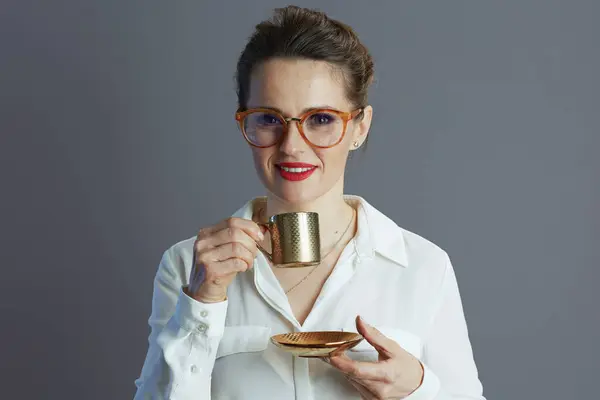 smiling 40 years old woman employee in white blouse with glasses and coffee cup against grey background.