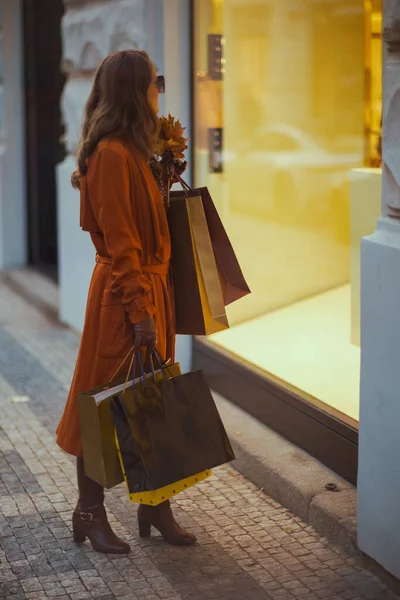 10月 こんにちは ショッピングバッグと秋の黄色い葉でオレンジ色のトレンチコートの若い女性のサイドビュー — ストック写真