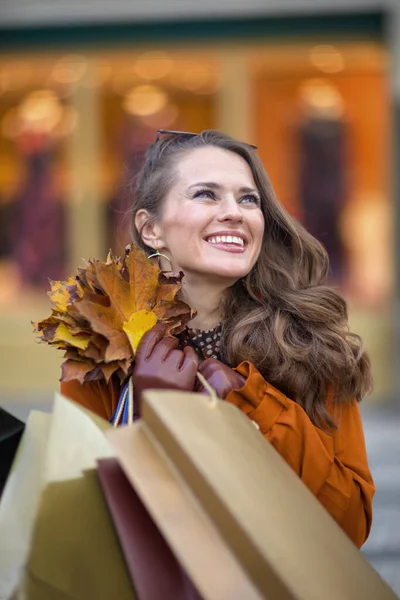Hello november. happy 40 years old woman in orange trench coat with shopping bags and autumn yellow leaves near store in the city.