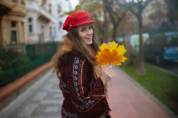 こんにちは秋 秋の葉と街を歩くスカーフで赤い帽子で幸せなトレンディな中年女性 — ストック写真