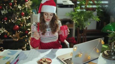 Noel zamanı. Noel Baba şapkalı mutlu iş kadını ve geleneksel kurabiyeli kırmızı Noel süveteri, bir fincan sıcak kakao ve dizüstü bilgisayar Noel ağacıyla modern yeşil ofiste şarkı söylüyor..