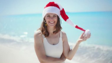 Gülümseyen orta yaşlı beyaz mayo giyen, uzun dalgalı saçlı, kumsalda çizgili Noel şapkalı bir kadın..