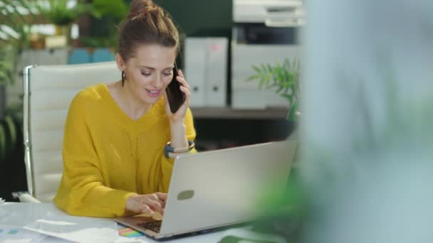 スマートフォンで話すラップトップと現代のグリーンオフィスで文書を扱う黄色いセーターで現代40歳の小さなビジネスオーナーの女性を微笑みます — ストック動画