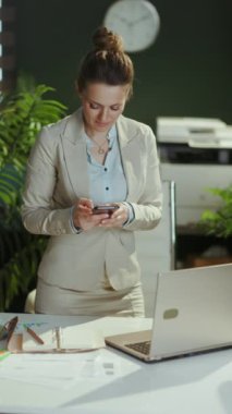 Modern yeşil ofisteki hafif takım elbiseli, akıllı telefon uygulaması kullanarak gülümseyen modern, küçük işletme sahibi kadın..