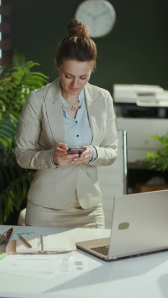 スマートフォンアプリを使用したラップトップと現代のグリーンオフィスの軽いビジネスススーツで現代の小さなビジネスオーナーの女性を微笑みます — ストック動画
