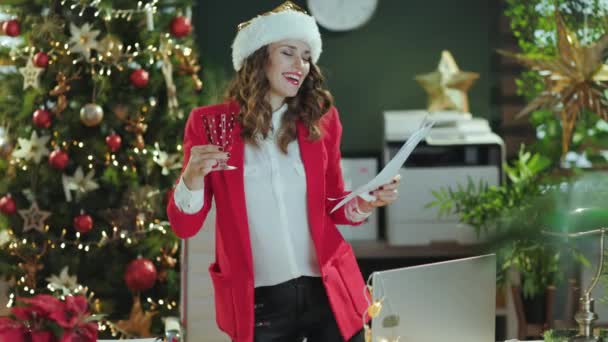クリスマス タイム ハッピーエレガントな小さなビジネスオーナーの女性サンタの帽子と赤いジャケット クリスマスツリーと現代グリーンオフィスのワイングラス — ストック動画