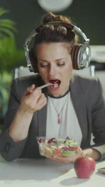 Sürdürülebilir iş yeri. Modern yeşil ofisteki gri takım elbiseli, kulaklıklı, salata yiyen mutlu modern muhasebeci kadın..