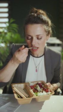 Sürdürülebilir iş yeri. Modern yeşil ofiste gri takım elbiseli mutlu bir modern kadın işçi salata yiyor..