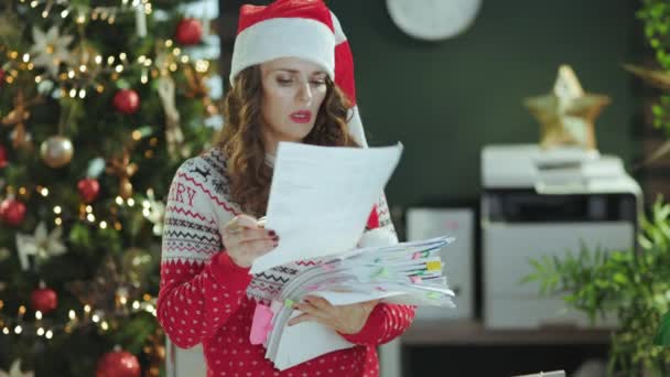 クリスマス タイム 疲れたスタイリッシュな中年ビジネスオーナー女性サンタ帽子と赤のクリスマスセーター クリスマスツリーで現代グリーンオフィスで働く文書 — ストック動画