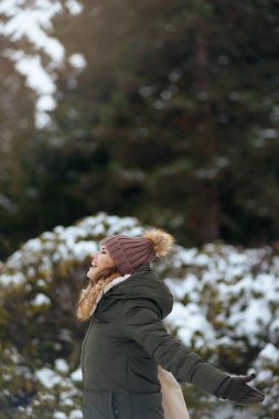 Gülümseyen, yeşil paltolu, kahverengi şapkalı modern kadın kışın şehir parkında bereli..