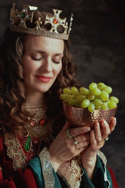 Kırmızı elbiseli ortaçağ kraliçesi. Üzüm tabağı ve tacı koyu gri arka planda..