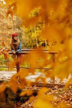 Merhaba sonbahar. 40 yaşında, kot tişörtlü, kırmızı şapkalı, güz yaprağı, atkısı ve çantası olan bir kadının tam boy portresi..