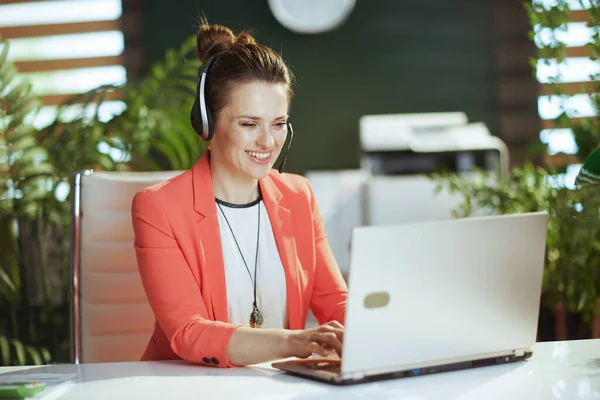 Sustainable workplace. smiling modern small business owner woman in modern green office in a red jacket with headset and laptop.