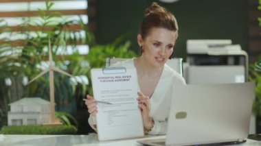 Devam etme zamanı. Beyaz bluzlu, modern yeşil ofisteki mutlu stil sahibi kadın emlakçı. Elinde pano ve konuşan video çağrısı var..