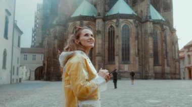 Prag Cumhuriyeti 'nde sarı bluzlu ve yağmurluklu gülümseyen genç bir kadın akıllı telefonları ve kulaklıkları ile şehir panoramasına karşı.