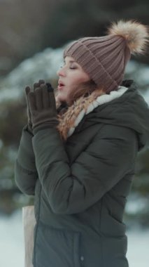 Kırk yıllık modern kadın yeşil ceketli ve kahverengi şapkalı kışın şehir parkında eldivenli ve bereli soğuk elleri nefesle ısıtan bir kadın..