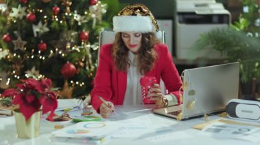 Noel zamanı. Noel Baba şapkası ve kırmızı ceketli mutlu modern iş kadını portresi, bir fincan sıcak içecek ve dizüstü bilgisayar modern yeşil ofiste Noel ağacı ile çalışıyor..