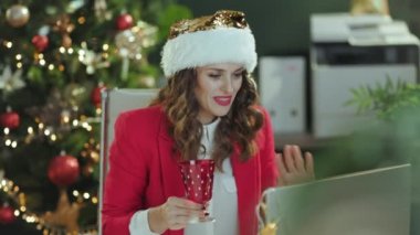 Noel zamanı. 40 yaşında, Noel Baba şapkalı, kırmızı ceketli, dizüstü bilgisayarı ve şarap kadehiyle modern yeşil ofisinde Noel ağacı olan bir iş kadını portresi..