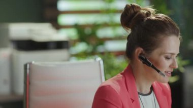 Sürdürülebilir iş yeri. Mutlu modern iş kadını iş yerinde kırmızı ceketli, kulaklıklı..
