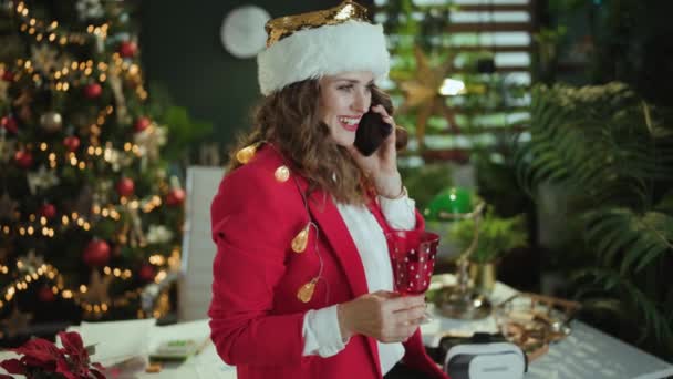 クリスマス タイム 笑顔のエレガントな小さなビジネスオーナーの女性 サンタ帽子と赤いジャケット クリスマスツリーと現代グリーンオフィスのスマートフォンで話すワイングラス — ストック動画