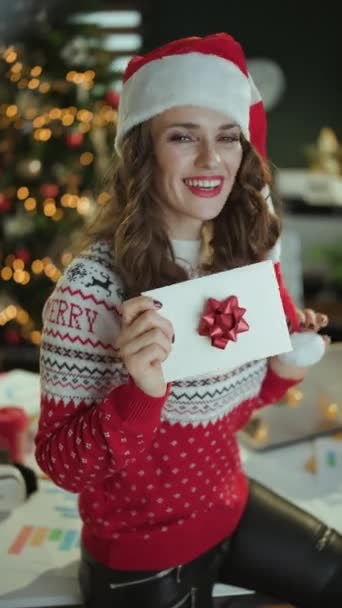 クリスマス タイム 笑顔の現代の中年ビジネスオーナーの女性サンタ帽子と赤のクリスマスセーター クリスマスツリーと現代グリーンオフィスの封筒 — ストック動画