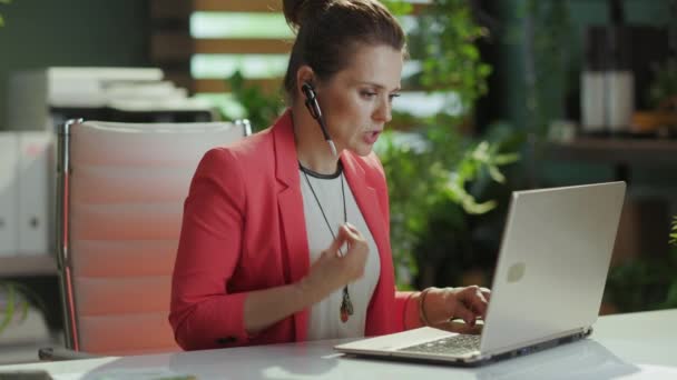 持続可能な職場 ヘッドセットとラップトップが付いている赤いジャケットで働く毛深い現代小さいビジネスオーナーの女性 — ストック動画