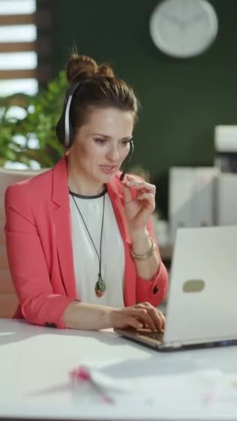 持続可能な職場 ヘッドセットとラップトップが付いている赤いジャケットの現代緑のオフィスの幸せな現代小企業の所有者の女性の肖像画 — ストック動画