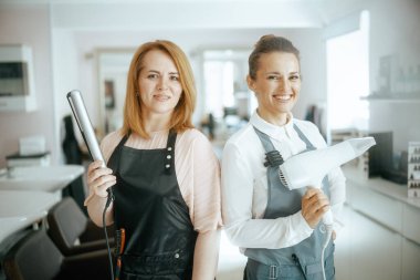 Modern güzellik stüdyosunda saç düzleştirici ve saç kurutma makinesiyle önlüklü iki orta yaşlı kadın kuaför kadının portresi..