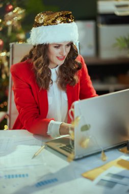 Noel zamanı. Noel Baba şapkalı, kırmızı ceketli, bilgisayarlı, modern yeşil ofiste Noel ağacıyla çalışan mutlu modern iş kadını..