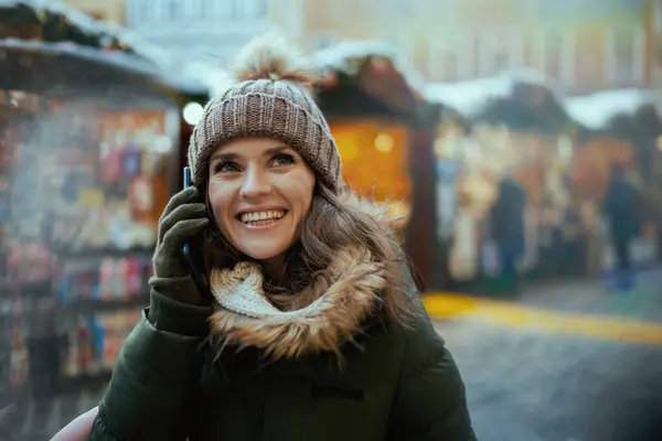 スマートフォンで話す市の冬のフェアで緑のコートと茶色の帽子で現代の女性を笑顔に — ストック写真