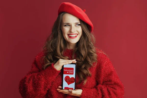 Happy Valentine. happy modern 40 years old woman with long wavy hair in red sweater and beret , smartphone and Valentine app.
