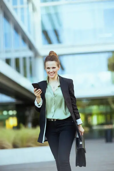 身穿黑色夹克 头戴智能手机 头戴公文包走路的40岁左右的现代女职员微笑着 — 图库照片