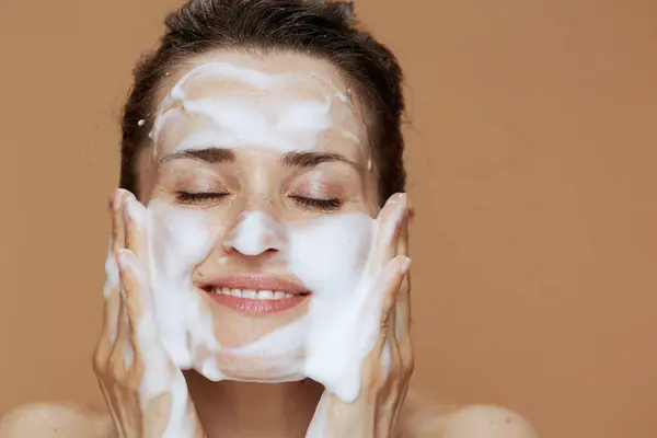 relaxed young woman with foaming face wash washing face against beige background.