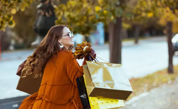こんにちは 市内のショッピングバッグと秋の黄色い葉とオレンジのトレンチコートのスタイリッシュな女性 — ストック写真