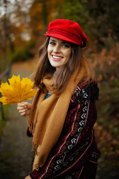 Hello autumn. happy stylish 40 years old woman in red hat with autumn leafs and scarf walking in the city park.