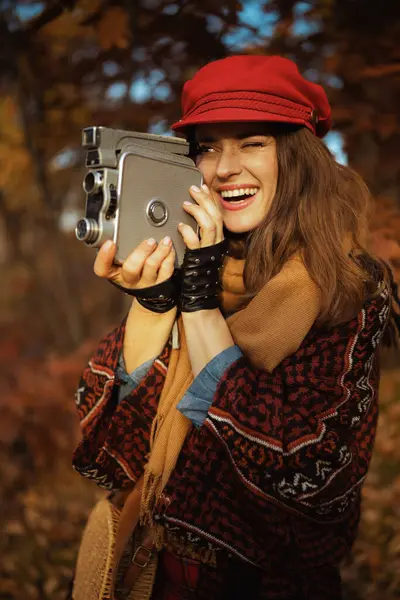 Hello autumn. smiling modern 40 years old woman in red hat with retro video camera, scarf and gloves in the city park.