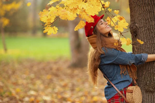 Hello autumn. relaxed modern woman in jeans shirt and red hat with autumn leafs, scarf and bag near tree in the city park.