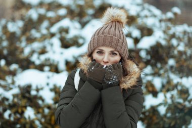 Mutlu, orta yaşlı, yeşil ceketli ve açık havada, şehir parkında kışın eldivenli ve bereli bir kadın. Soğuk elleri, karlı dalların yanında ısıtan bir şapka..