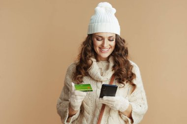 Merhaba kış. Bej kazaklı, eldivenli ve şapkalı, akıllı telefon ve kredi kartı ile izole edilmiş, e-ticaret sitesinden online alışveriş yapan zarif bir kadın..
