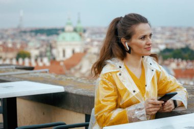 Prag Cumhuriyeti 'nde sarı bluz ve yağmurluk giyen, kulaklıklı, şehir panoramasına karşı akıllı telefon kullanan, son moda yalnız gezgin kadın..